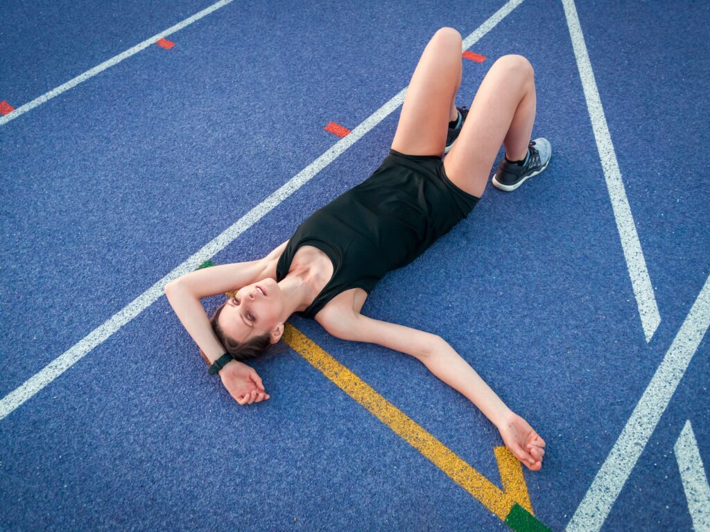 Eine erschöpfte Läuferin liegt nach einem Training auf einer blauen Bahn und ruht sich aus.
